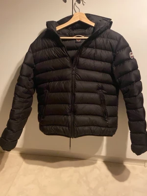 Svart Colmar pufferjacka med huva - Svart pufferjacka från Colmar med huva och dragkedja framtill. Jackan har två sidofickor med dragkedja och ett broderat Colmar-märke på ärmen. Den är quiltad och har en normal passform, perfekt för kyliga dagar. Pris är inte hugget i sten