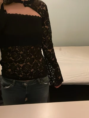Svart spetsig bolerotopp med rosett - Säljer en svart bolerotopp från Nelly med långärmade spetsärmar .Toppen har ett  bandeau-liknande bröstparti och genomskinliga blommiga spetsdetaljer. Dör för denna tröja perfekt till fest dock beställde jag fel strlk. Ny lappen är kvar dessutom slutsåld☺️