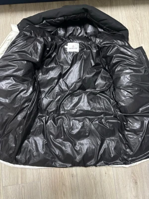 Vit Moncler pufferjacka med svart huva - Säljer en fet pufferjacka från Moncler i vit med svart huva och insida. Jackan har Moncler-logga på ärmen, stora fickor och dragkedja med tryckknappar. Materialet är glansig polyester och dunfyllning, perfekt för kalla dagar.