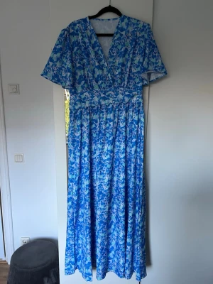 Blommig blå långklänning XL - Säljer en långklänning med blommigt mönster i olika blå och ljusa toner. Klänningen har v-ringad hals, korta ärmar och ett luftigt fall. Från SHEIN.