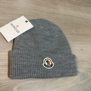 Moncler mössa  - Snygg grå ribbstickad mössa från Moncler med klassisk uppvikt kant och broderad logotyp framtill. Tillverkad i mjukt material som håller dig varm och ger en clean look. Perfekt accessoar för kyliga dagar.