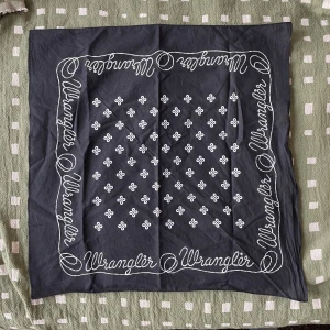 Svart bandana från Wrangler - Svart halsduk/bandana från Wrangler med vita små blommiga prickmönster och logotypen tryckt längs kanterna i repstil. Fyrkantig form, tunn och lätt, perfekt att knyta runt halsen eller huvudet. Materialet är mjuk bomull. Helt ny!