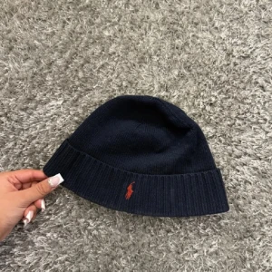 Mörkblå stickad mössa Polo Ralph Lauren - Snygg mörkblå stickad mössa från Polo Ralph Lauren med klassisk röd logga broderad framtill. Mössan har uppvikt kant och är tillverkad i mjukt stickat material som håller dig varm och stilren under kyliga dagar.