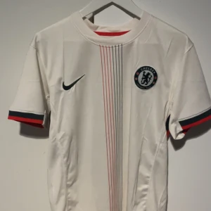 Chelsea Colwill #6 vit fotbollströja - Chelsea FC Colwill #6 matchtröja från Nike i vit polyester med tunna röda och svarta ränder på framsidan. Kortärmad modell med klubbmärke och Nike-logga på bröstet. Ärmsluten har svarta och röda detaljer för extra stil. REPLIKA!