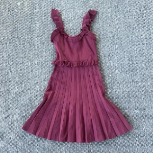 Vinröd stickad plisserad klänning från Zara - Supersöt vinröd klänning från Zara med plisserad kjol och volangdetaljer vid axelbanden och midjan. Ärmlös modell med snyggt fall och ribbad struktur i tyget. Perfekt för dig som vill sticka ut med en feminin och trendig look.