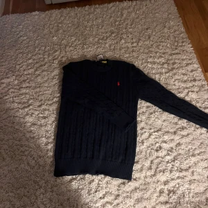 Mörkblå stickad tröja Polo Ralph Lauren - Snygg mörkblå stickad tröja från Polo Ralph Lauren med klassiskt kabelstickat mönster och röd broderad logga på bröstet. Rund hals och långa ärmar, perfekt för dig som gillar stilrena och tidlösa plagg.
