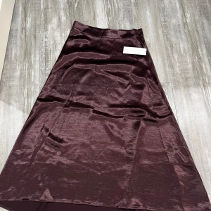 Mörklila långkjol i satin från Gina Tricot - Supersnygg mörklila långkjol från Gina Tricot i glansigt satinmaterial. Kjolen har en klassisk A-linjeform och slät midja, perfekt för dig som vill ha en elegant och trendig look. Passar dig som gillar stilrena plagg med lite extra shine.