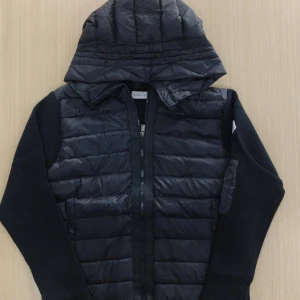 Svart Moncler pufferjacka med huva - Svart pufferjacka från Moncler med quiltad framsida och huva. Jackan har stickade ärmar och detaljer som dragkedja framtill och ficka med dragkedja på ärmen. Perfekt för dig som vill ha en snygg och sportig look med exklusiv känsla.