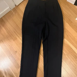 Svarta kostymbyxor från NA-KD - Snygga svarta kostymbyxor från NA-KD med klassisk rak passform och hög midja. Byxorna har pressveck framtill och är perfekta för en clean och stilren look. Materialet känns mjukt och har en lätt stretch för extra komfort.