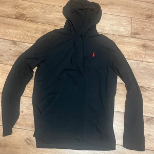 Svart hoodie från Polo Ralph Lauren - Svart hoodie från Polo Ralph Lauren med röd broderad logga på bröstet. Tröjan har huva med snörning och långa ärmar. Tillverkad i mjuk bomull som är skön att bära. Perfekt för chill dagar och enkel att matcha med jeans eller joggers.