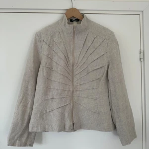 Beige linnéjacka från Robell - Snygg beige jacka från Robell i linne med coola veckade detaljer framtill och baktill som ger en unik look. Jackan har hög krage, dragkedja och lång ärm. Perfekt för dig som gillar stilrena och annorlunda plagg.