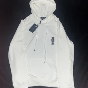  Polo Ralph Lauren Hoodie - Vit hoodie från Polo Ralph Lauren med broderad nalle på bröstet. Tröjan har huva med snörning, känguruficka och custom fit-passning. Perfekt för dig som gillar stilrena och klassiska plagg med en twist.