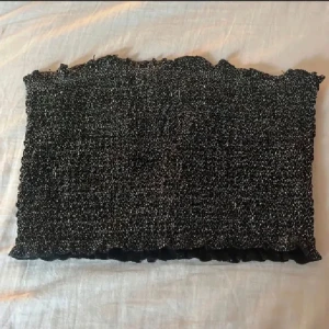 Glittrig svart  tubtopp - Säljer en glittrig svart  tubtopp med silvriga detaljer. Toppen är stretchig och har en vågig kant upptill och nedtill för en cool look. Perfekt att styla med jeans eller kjol för att sticka ut lite extra. Materialet är syntet och toppen är axelbandslös. Perfekta toppen till nyår!
