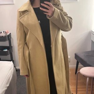 Beige lång kappa med bälte - Säljer en beige kappa med snygg krage och bälte i midjan. Jackan är lång, har slits bak och stora fickor på sidorna. Materialet är mjukt och har en diskret struktur. Perfekt för dig som vill ha en clean och chill look under höst och vinter. Använd endast en gång 