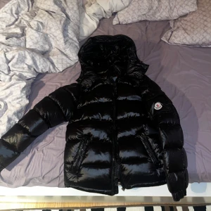 Moncler Maya - Snygg och varm Moncler Maya i svart – en riktig vinterfavorit. Jackan är fylld med vitt ankdun (ca 90 % dun), vilket ger utmärkt volym och värme under kalla dagar.   Detaljer: • Storlek: passar S–M (motsvarar ungefär EU 50/M i originalmärkning) • Färg: svart • Material: glansig nylon • Fyllning: vitt ankdun med hög fyllnadsgrad • Huvan är avtagbar • Stängning: dragkedja • Passform: normal • Modellår: vinter 2024 • Jackan har fungerande NFS (äkhetsverifiering) En bekväm, stilren och riktigt varm 
