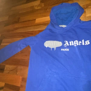 Blå hoodie från Palm Angels - Säljer en blå hoodie från Palm Angels med vit logga och texten 'Paris' på bröstet. Tröjan har huva, lång ärm och en klassisk känguruficka framtill. Materialet är mjukt och känns skönt mot huden, perfekt för chill dagar. Passformen är normal.