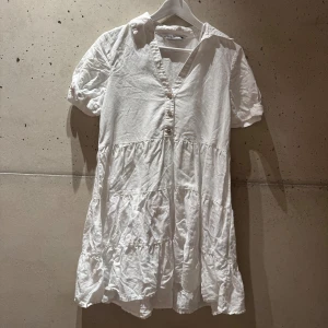 Vit kort klänning från Zara i linne - Fräsch vit klänning från Zara i linne med korta puffärmar, v-ringning och knappar framtill. Modellen är kort med volangdetaljer och har en avslappnad passform. Perfekt för varma dagar och en chill stil. Fin också till studenten för en minimalistisk look! Använt fåtal gånger men skrynklig pga varit vikt i garderoben!