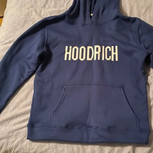 Blå HOODRICH hoodie med tryck - Snygg blå hoodie från HOODRICH med vit logga tryckt på bröstet. Klassisk känguruficka framtill och huva. Perfekt för dig som gillar streetwear och vill ha en stilren look.
