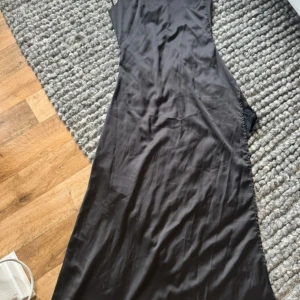 Svart långklänning med spets från CBR - Svart långklänning från CBR i storlek S. Klänningen har en elegant v-ringning och ärmlös design med snygga spetsdetaljer på sidorna. Materialet är mjukt och har en satinliknande finish som ger en lyxig känsla. Perfekt för dig som vill ha en stilren och sofistikerad look.