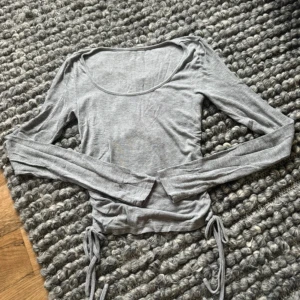Grå långärmad topp med snörning - Säljer en grå långärmad topp med rund halsringning och snygga snörningsdetaljer i sidorna. Toppen är croppad och har en slim passform som framhäver figuren. Perfekt att styla med jeans eller kjol för en trendig look.