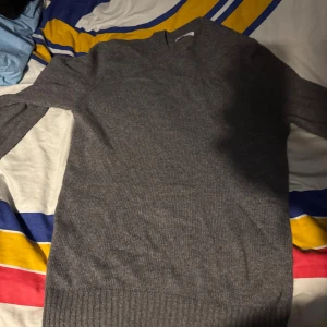 Grå stickad tröja med rund hals - En enkel och stilren grå stickad tröja med rund hals och långa ärmar. Tröjan har ribbade muddar vid ärmslut och nederkant, vilket ger en snygg och klassisk look. Perfekt för att hålla sig varm under kyliga dagar.