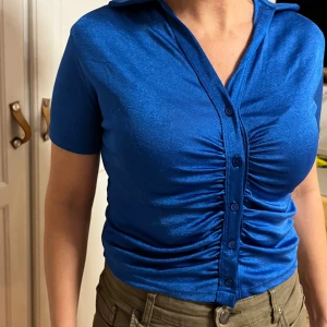 Blå kortärmad skjorta från Primark - Snygg blå skjorta från Primark i storlek S. Skjortan har korta ärmar, knappar framtill och cool rynkad detalj längs knappraden. Materialet är glansigt och känns mjukt mot huden, perfekt för dig som gillar att sticka ut med färg. Passformen är tight och kragen ger en klassisk touch.