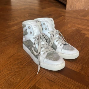 Zadig & Voltaire skor  - Säljer ett par vita high-top sneakers från Zadig & Voltaire med glittriga silverdetaljer och coola snören med text. Skorna har snörning, dragkedja på sidan och är köpta i butiken i Paris. Jätte fint skick tyvärr bara lite förstora för mig❤️