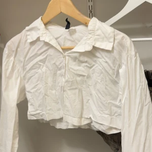 Vit croppad skjorta från H&M Divided - Vit croppad skjorta från H&M Divided med klassisk krage och långa ärmar. Skjortan är i bomull och har en loose fit som ger en avslappnad vibe. Perfekt att styla med höga jeans eller kjol för en trendig look.