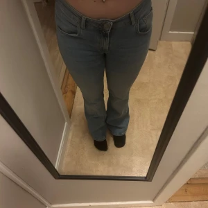 Bootcut ljusblå jeans - Säljer ett par ljusblå bootcut jeans med klassisk femficksdesign och bälteshällor. Jeansen har en låg midja och är tillverkade i mjukt denimtyg som sitter snyggt över höfterna och låren innan de blir vidare nedtill.