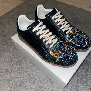 Maison Margiela gats - Maison Margiela gats – EU 44 (svart/multicolor) Stilrena Margiela-sneakers i svart läder med färgstänks-design och klassisk gummisula. Vita snören och vit insida. Passar både till jeans och dressat.  Skick: aldrig använda, endast testade inomhus. Se bilder för detaljer.  Info 	•	Storlek: EU 44 	•	Färg: Svart med multicolor-färgstänk 	•	Material: Läder med gummisula  Om köp & pris Köpta på Vinted. Säljaren uppgav att de var äkta, men vid köp via begagnade plattformar kan man aldrig vara 100 %