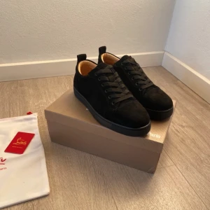Svarta sneakers från Christian Louboutin - Stilrena svarta sneakers från Christian Louboutin i mocka med klassisk röd sula och svarta snören. Insidan är ljusbrun och skon har en rund tå. Perfekta för dig som vill ha en lyxig och tidlös look med en ikonisk detalj.
