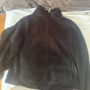 Svart teddyjacka med dragkedja - Mysig svart teddyjacka med hel dragkedja framtill och hög krage. Jackan har en fluffig och mjuk yta som ger en chill vibe och passar perfekt när du vill hålla dig varm. Enkel och stilren design utan synliga detaljer eller mönster.