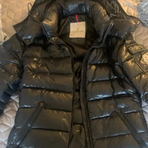Svart Moncler pufferjacka med huva - Säljer en svart pufferjacka från Moncler med glansig finish och huva. Jackan har Moncler-logga på ärmen, praktiska fickor med dragkedja och en cool quiltad design. Perfekt för dig som vill ha en varm och snygg jacka med streetkänsla. NI FÅR JÄTTE GÄRNA SKRIVA VA PRIS FÖRSLAG🤗