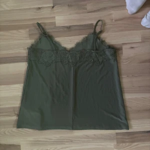 Mörkgrön linnetopp med spets - Snygg mörkgrön linnetopp med tunna axelband och v-ringad hals. Toppen har dekorativ spets längs överdelen som ger en extra detalj. Materialet är mjukt och lätt, perfekt för varma dagar. Enkel och stilren design som är lätt att matcha med jeans eller kjol.