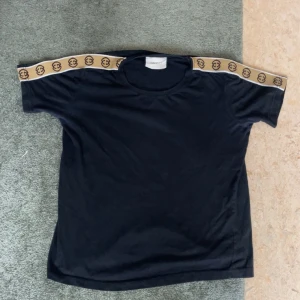 Svart t-shirt med logoband från Select - Svart t-shirt från Select med korta ärmar och beige band med logomönster längs axlarna. Rund halsringning och rak passform. Materialet känns mjukt och lätt, perfekt för en chill look. Snygg detalj med logobandet som ger extra edge.