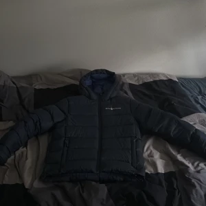 Svart Sail Racing pufferjacka med huva - Säljer en svart pufferjacka från Sail Racing med luva och blått foder. Jackan har dragkedja framtill, två sidofickor med dragkedja och logga på bröstet. Perfekt för kalla dagar och har en schysst, sportig look. Passar dig som gillar stilren design och funktion.