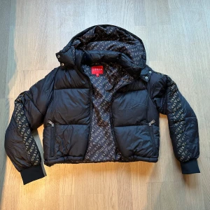 Svart pufferjacka från Guess  - Svart pufferjacka från Guess med logotypmönstrat foder och detaljer på ärmarna. Jackan har huva, dragkedja framtill och två fickor med zip. Croppad passform och breda muddar vid ärmslut. Perfekt för dig som vill ha en snygg och varm jacka med street vibe.