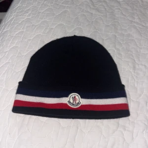 Moncler mössa med randig kant - Svart Moncler mössa i mjuk ull med klassisk uppvikt kant. Kanten har breda ränder i marinblått, vitt och rött samt Moncler-logga framtill. Perfekt accessoar för att hålla värmen och ge en sportig touch till din outfit.