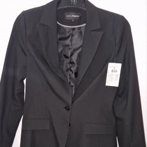 Svart kavaj från KappAhl, strl 34 - Stilren svart kavaj från KappAhl med klassisk krage och två knappar framtill. Fodrad insida i blankt tyg och fickdetaljer fram. Långa ärmar med knappar vid ärmslut. Perfekt för en snygg och clean look.