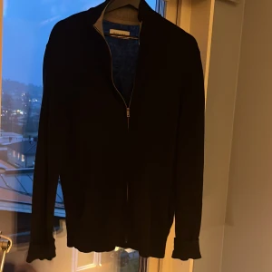 Svart kofta med dragkedja Pier One - Svart kofta från Pier One med stilren design och hel dragkedja framtill. Tröjan har långärmade ärmar och ribbad krage i grått. Perfekt för dig som gillar klassiska och enkla plagg med snygg passform.