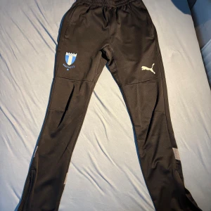 Svarta Malmö FF träningsbyxor Puma XS - Svarta träningsbyxor från Puma i storlek XS med Malmö FF-logga på ena benet och Puma-logga på andra. Byxorna har elastisk midja, smal passform och ventilerande detaljer vid vaden. Perfekta för fotboll eller träning.