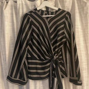 Svart randig omlottblus med knyt - Säljer en svart blus med vita och beige ränder, omlottmodell med snygg knyt framtill. Blusen har långa ärmar med breda manschetter och ett coolt mönster som ger en modern vibe. Perfekt för dig som gillar stilrena detaljer och vill sticka ut.