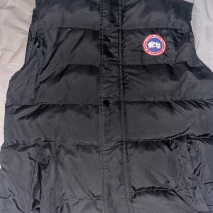 Svart dunväst från Canada Goose  - Svart dunväst från Canada Goose med klassisk logga på bröstet och broderad Canada Goose-text på ryggen. Västen är quiltad och har praktiska fickor framtill. Perfekt för lager-på-lager under kyliga dagar.  Storlek S men passar också M. Kom om för mer bilder. Fickorna är lite skadade men inget man tänker på 