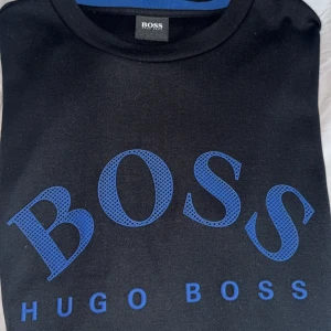 Svart sweatshirt från Hugo Boss - Svart sweatshirt från Hugo Boss med stort blått BOSS-tryck framtill och rund halsringning. Tröjan har långa ärmar och en klassisk passform. Perfekt för dig som gillar stilrena och snygga märkesplagg.