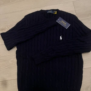 Mörkblå stickad tröja Polo Ralph Lauren - Snygg mörkblå stickad tröja från Polo Ralph Lauren med klassisk kabelstickning och vit broderad logga på bröstet. Rund hals och långa ärmar med ribbade muddar. Perfekt för dig som gillar stilrena och tidlösa plagg. passar mer som en liten