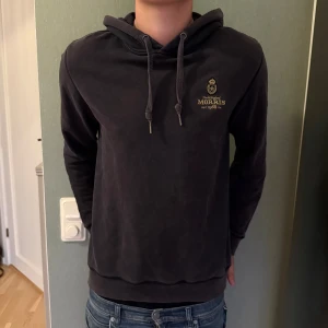 Mörkblå hoodie från Morris - Mörkblå hoodie från Morris med guldfärgat broderat logotyptryck på bröstet. Tröjan har luva med snörning. Perfekt för chill dagar och enkel att matcha med jeans. Fint skick men något urtvättad. 