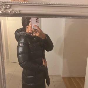 Svart lång jacka från Colmar - Säljer en svart, lång och glansig dunjacka från Colmar med huva och dragkedja. Jackan har en quiltad design och logotyp på ärmen. Perfekt för kalla dagar när du vill hålla stilen och värmen.