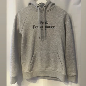 Grå hoodie från Peak Performance - Snygg grå hoodie från Peak Performance med svart broderad logga på bröstet. Tröjan har huva med snörning, känguruficka och ribbade muddar. Perfekt för chill dagar eller när du vill ha en enkel och stilren look. Den är använt få tal gånger (2-3 ggr). Ny pris 1300 kr