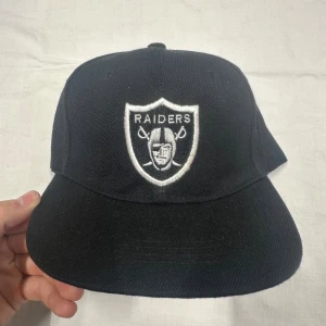 Svart Raiders keps med broderad logga - Snygg svart keps med broderad Raiders-logga i vitt framtill. Klassisk snapback-modell med justerbar plastrem bak. Kepsen har rak skärm och är tillverkad i bomullstyg, perfekt för dig som gillar sportig streetstyle.