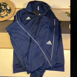 Blå Adidas träningsset i medium - Snyggt blått träningsset från Adidas i storlek medium. Setet består av en zip-jacka med ståkrage och matchande byxor med vita detaljer längs sidorna. Båda delarna har Adidas-loggan tryckt i vitt. Perfekt för dig som gillar sportig stil.